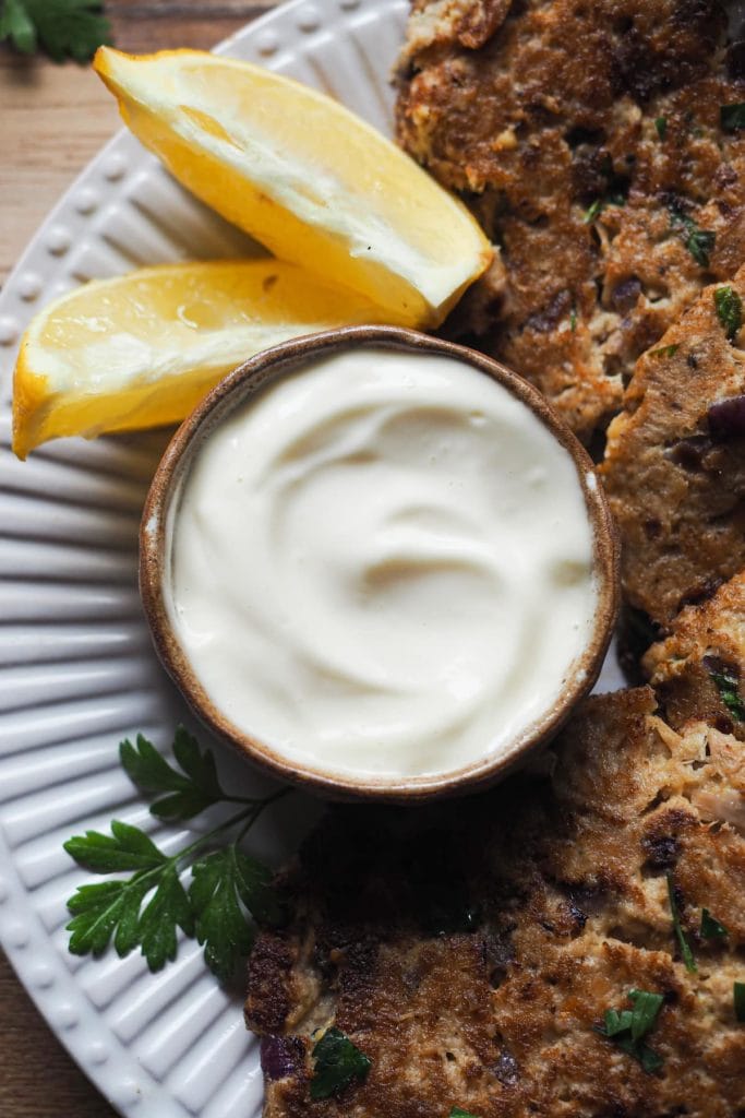 Esta receta de Hamburguesas de Atún es deliciosa, sabrosa y te salva las cenas de entre semana. Están hechas con ingredientes que siempre solemos tener en casa y también son perfectas para preparar comida con antelación! Además, es una receta sin gluten. Esta es una foto de la mayonesa que se sirve con las hamburguesas. Está en un bol pequeño rodeada con perejil, hamburguesas de atún y un par de rodajas de limón.