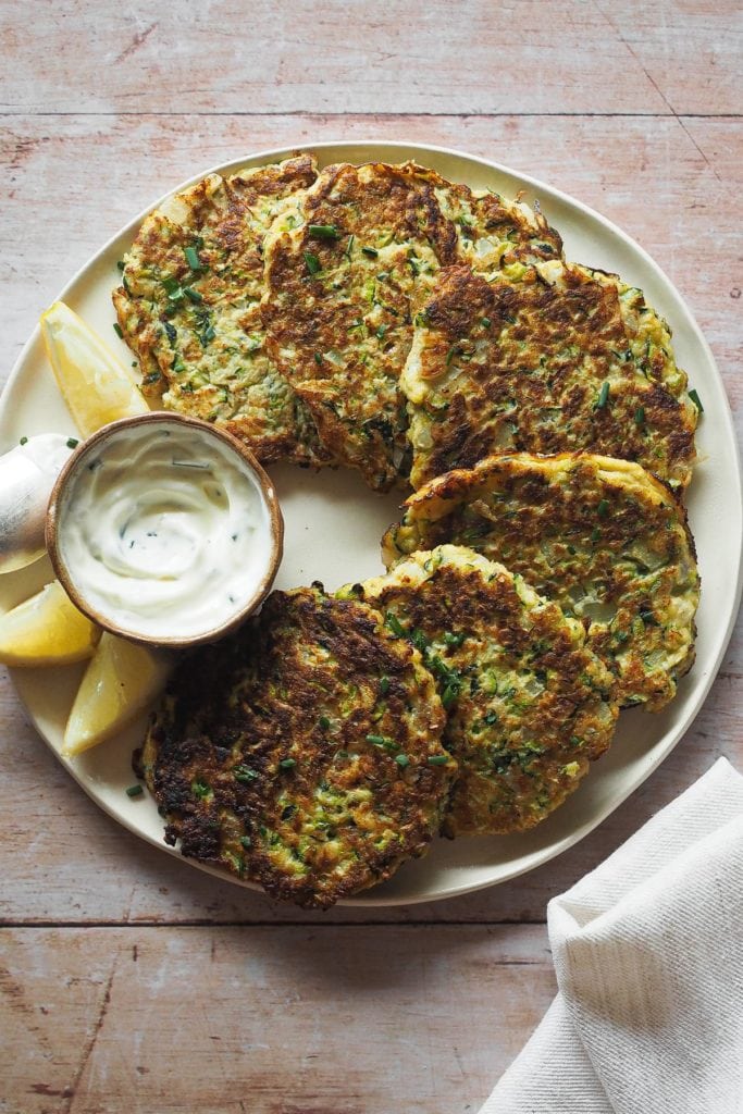 Estas Tortitas de Calabacín con Crème Fraîche al Limón y Hierbas son fenomenales para las cenas entre semana. No contienen gluten y combinan huevos, calabacín y una mezcla de hierbas y especias que las hacen tan deliciosas como ligeras. En esta foto cenital se ven las tortitas en un plato, dispuestas en semicírculo rodeando un pequeño bol con el dip de crème fraîche. A bajo a la derecha se ve una puntita de un trapo de cocina color hueso.