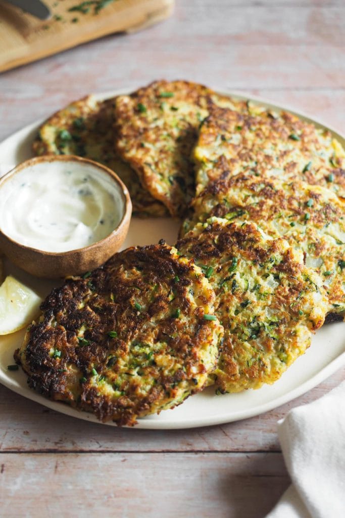 Estas Tortitas de Calabacín con Crème Fraîche al Limón y Hierbas son fenomenales para las cenas entre semana. No contienen gluten y combinan huevos, calabacín y una mezcla de hierbas y especias que las hacen tan deliciosas como ligeras. La foto está tomada con un ángulo de unos 45 grados y se ven las tortitas en un plato, dispuestas en semicírculo rodeando un pequeño bol con el dip de crème fraîche. Al fondo, en la esquina izquierda, se ve la punta de una tabla de madera con cebollino picado y la punta de un cuchillo. A bajo a la derecha se ve una puntita de un trapo de cocina color hueso.