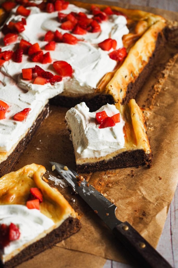 Brownie Cheesecake con Nata y Fresas. Abajo, un brownie denso y jugoso. Luego, una capa de cheesecake de vainilla y para terminar, nata montada y fresas. Y encima es fácil de hacer y (casi) imposible de liarla, te lo prometo! En la foto se ve un trozo cortado de la tarta en el que se distinguen muy claramente las 3 capas: la parte de abajo es un brownie jugoso y encima lleva el cheesecake y la nata con las fresas troceadas y bañadas con su propio sirope.