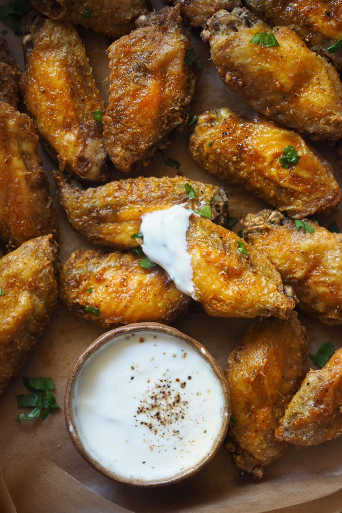 Alitas de pollo crujientes al horno adictivas. Llevan un aliño de ajo y pimentón que es, literal, para chuparse los dedos y van acompañadas de un dip de yogur y limón que balancea todos los sabores. Aquí se ven las alitas con el dip servido en un bol y una de las alitas con la punta embadurnada de dip.