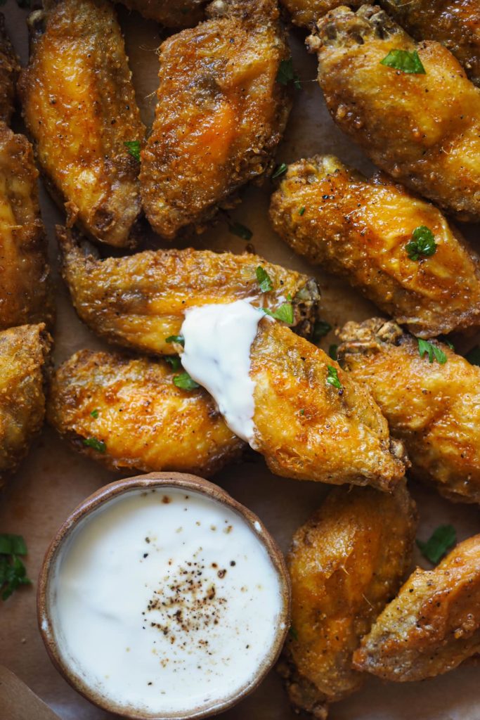 Alitas de pollo crujientes al horno adictivas. Llevan un aliño de ajo y pimentón que es, literal, para chuparse los dedos y van acompañadas de un dip de yogur y limón que balancea todos los sabores. Aquí se ven las alitas con el dip servido en un bol y una de las alitas con la punta embadurnada de dip.