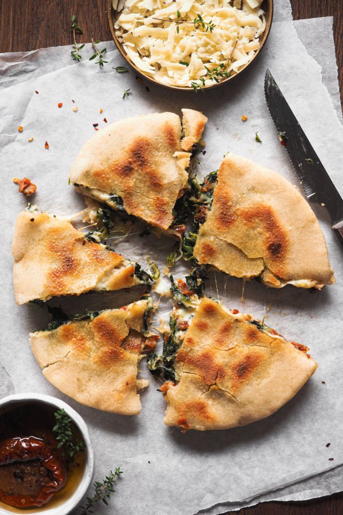 Sandwich de Queso Espinacas y Tomate Seco con Pita (o Quesapita de Espinacas y Tomate Seco). Sencillo y a la vez bestial. En lugar de usar pan clásico o de molde, se hace con pita. Este se rellena con una mezcla de distintos quesos, espinacas y tomate seco y de aquí a la sartén para que el queso quede súper derretidito y el pita crujiente. Vamos, un sandwich perfecto para cualquier día de la semana, a cualquier hora, ¡que te va a encantar! En la foto cenital se ve una quesapita cortada en triángulos, cada uno como una porción. Arriba hay un pequeño plato con más queso, arriba a la derecha un cuchillo de sierra y abajo a la izquierda un pequeño bol con más tomate seco.