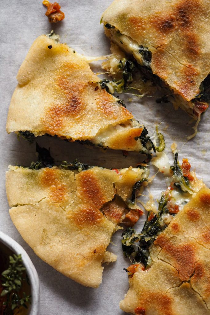 Sandwich de Queso Espinacas y Tomate Seco con Pita (o Quesapita de Espinacas y Tomate Seco). Sencillo y a la vez bestial. En lugar de usar pan clásico o de molde, se hace con pita. Este se rellena con una mezcla de distintos quesos, espinacas y tomate seco y de aquí a la sartén para que el queso quede súper derretidito y el pita crujiente. Vamos, un sandwich perfecto para cualquier día de la semana, a cualquier hora, ¡que te va a encantar! En la foto cenital se ve la mitad de una quesapita cortada en triángulos a modo porción.