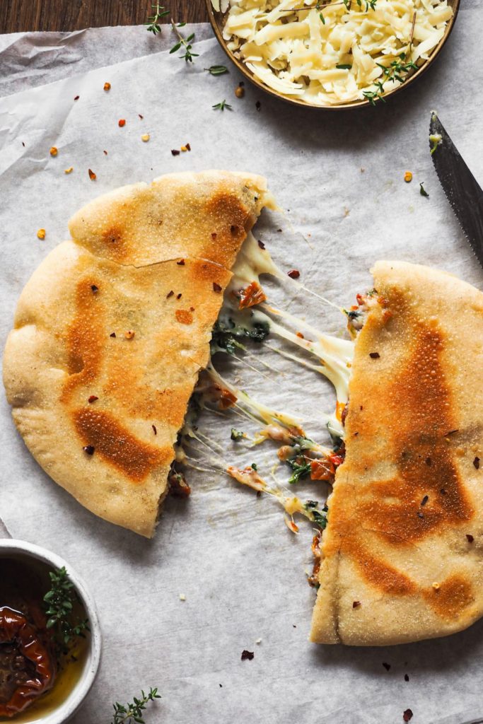 Sandwich de Queso Espinacas y Tomate Seco con Pita (o Quesapita de Espinacas y Tomate Seco). Sencillo y a la vez bestial. En lugar de usar pan clásico o de molde, se hace con pita. Este se rellena con una mezcla de distintos quesos, espinacas y tomate seco y de aquí a la sartén para que el queso quede súper derretidito y el pita crujiente. Vamos, un sandwich perfecto para cualquier día de la semana, a cualquier hora, ¡que te va a encantar! En la foto cenital se ve una quesapita cortada en 2 mitades separadas con queso derretido entre ellas a modo de filamentos, unos más gruesos y otros más delgados. Arriba hay un pequeño plato con más queso, arriba a la derecha un cuchillo de sierra y abajo a la izquierda un pequeño bol con más tomate seco.