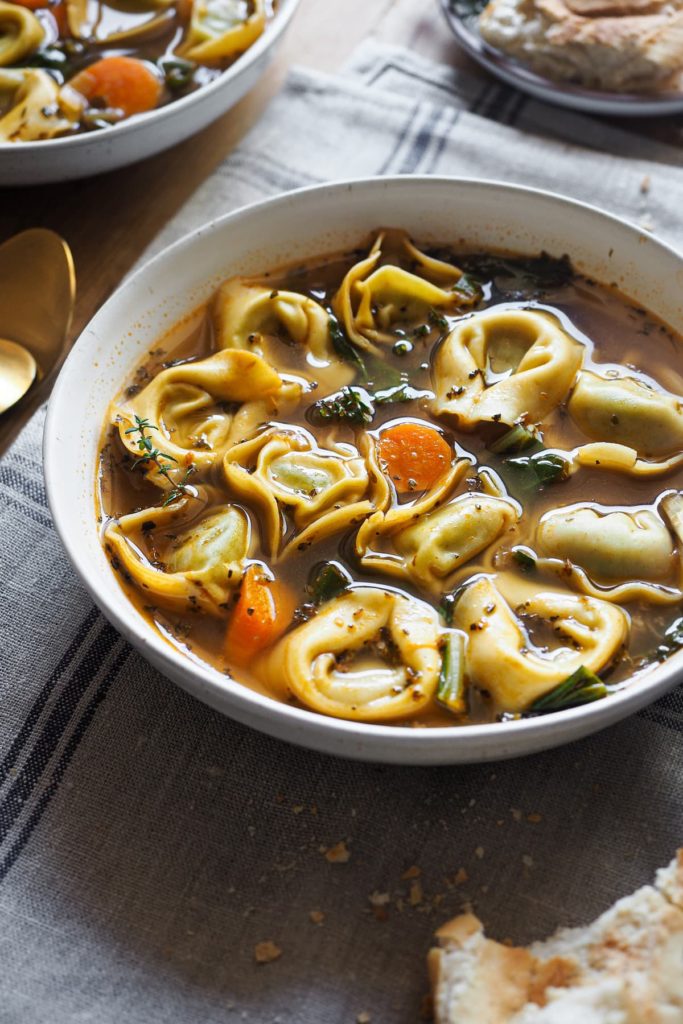 ¡En una noche fría, esta receta de Sopa de Tortellini Saludable en 30 minutos será tu mejor amiga! Está repleta de verduras y pasta, es reconfortante, sabrosa y fácil de preparar. Si estás en busca de comidas nutritivas y calentitas, te encantará esta sopa de tortellini. Se hace en poco más de 30 minutos, por lo que es fácil de preparar entre semana. Los tortellini hacen que llene y que la sopa entre más por los ojos, por lo que la hace ideal para niños. En esta foto a unos 30º se ve la sopa servida en un plato con tortellini, acelgas, zanahoria, el caldo y un poco de tomillo fresco como decoración. Hay un poco de pan al lado y el plato está encima de un paño gris.