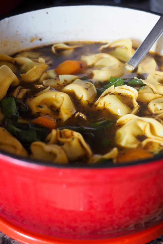 ¡En una noche fría, esta receta de Sopa de Tortellini Saludable en 30 minutos será tu mejor amiga! Está repleta de verduras y pasta, es reconfortante, sabrosa y fácil de preparar. Si estás en busca de comidas nutritivas y calentitas, te encantará esta sopa de tortellini. Se hace en poco más de 30 minutos, por lo que es fácil de preparar entre semana. Los tortellini hacen que llene y que la sopa entre más por los ojos, por lo que la hace ideal para niños. En esta foto a unos 30º se ve la sopa en la olla en la que se ha cocinado con un cucharón dentro.