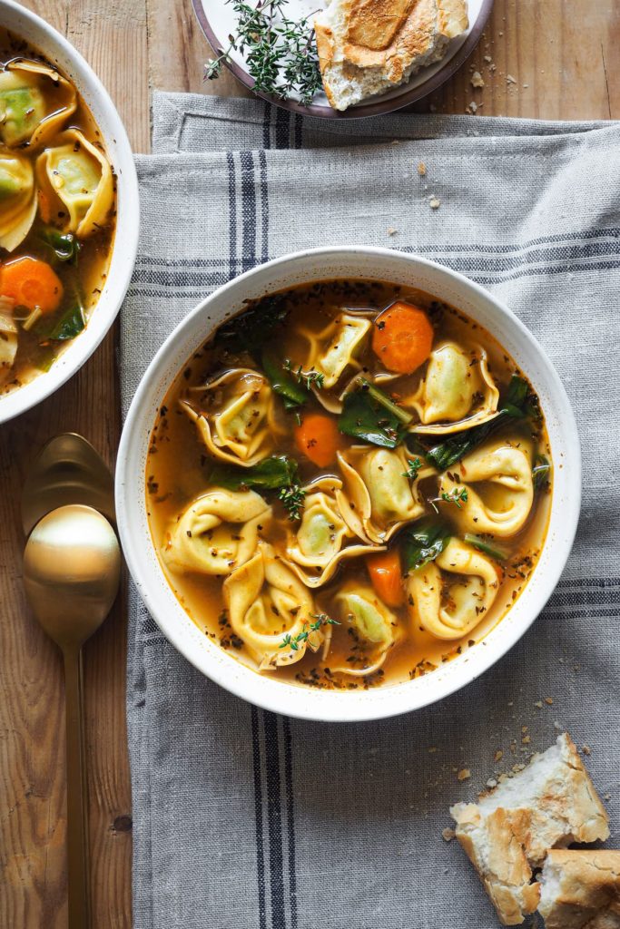 ¡En una noche fría, esta receta de Sopa de Tortellini Saludable en 30 minutos será tu mejor amiga! Está repleta de verduras y pasta, es reconfortante, sabrosa y fácil de preparar. Si estás en busca de comidas nutritivas y calentitas, te encantará esta sopa de tortellini. Se hace en poco más de 30 minutos, por lo que es fácil de preparar entre semana. Los tortellini hacen que llene y que la sopa entre más por los ojos, por lo que la hace ideal para niños. En esta foto cenital se ve la sopa servida en un plato con tortellini, acelgas, zanahoria, el caldo y un poco de tomillo fresco como decoración. Hay un poco de pan al lado y el plato está encima de un paño gris.