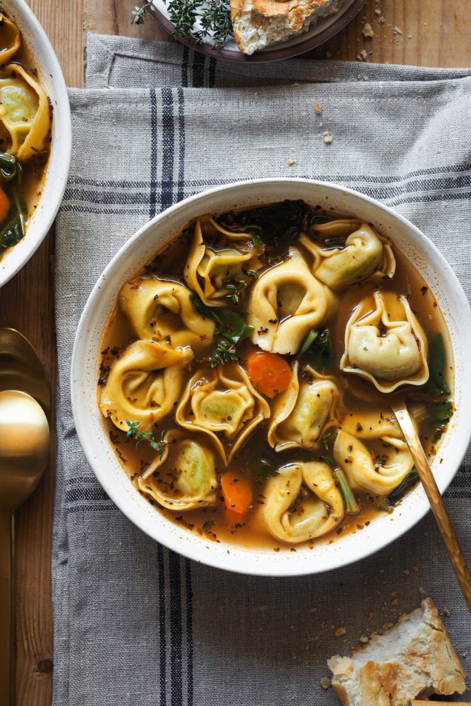 ¡En una noche fría, esta receta de Sopa de Tortellini Saludable en 30 minutos será tu mejor amiga! Está repleta de verduras y pasta, es reconfortante, sabrosa y fácil de preparar. Si estás en busca de comidas nutritivas y calentitas, te encantará esta sopa de tortellini. Se hace en poco más de 30 minutos, por lo que es fácil de preparar entre semana. Los tortellini hacen que llene y que la sopa entre más por los ojos, por lo que la hace ideal para niños. En esta foto cenital se ve la sopa servida en un plato con tortellini, acelgas, zanahoria, el caldo y un poco de tomillo fresco como decoración. Dentro del plato hay una cuchara con un tortellini y al lado hay un poco de pan. El plato está encima de un paño gris.