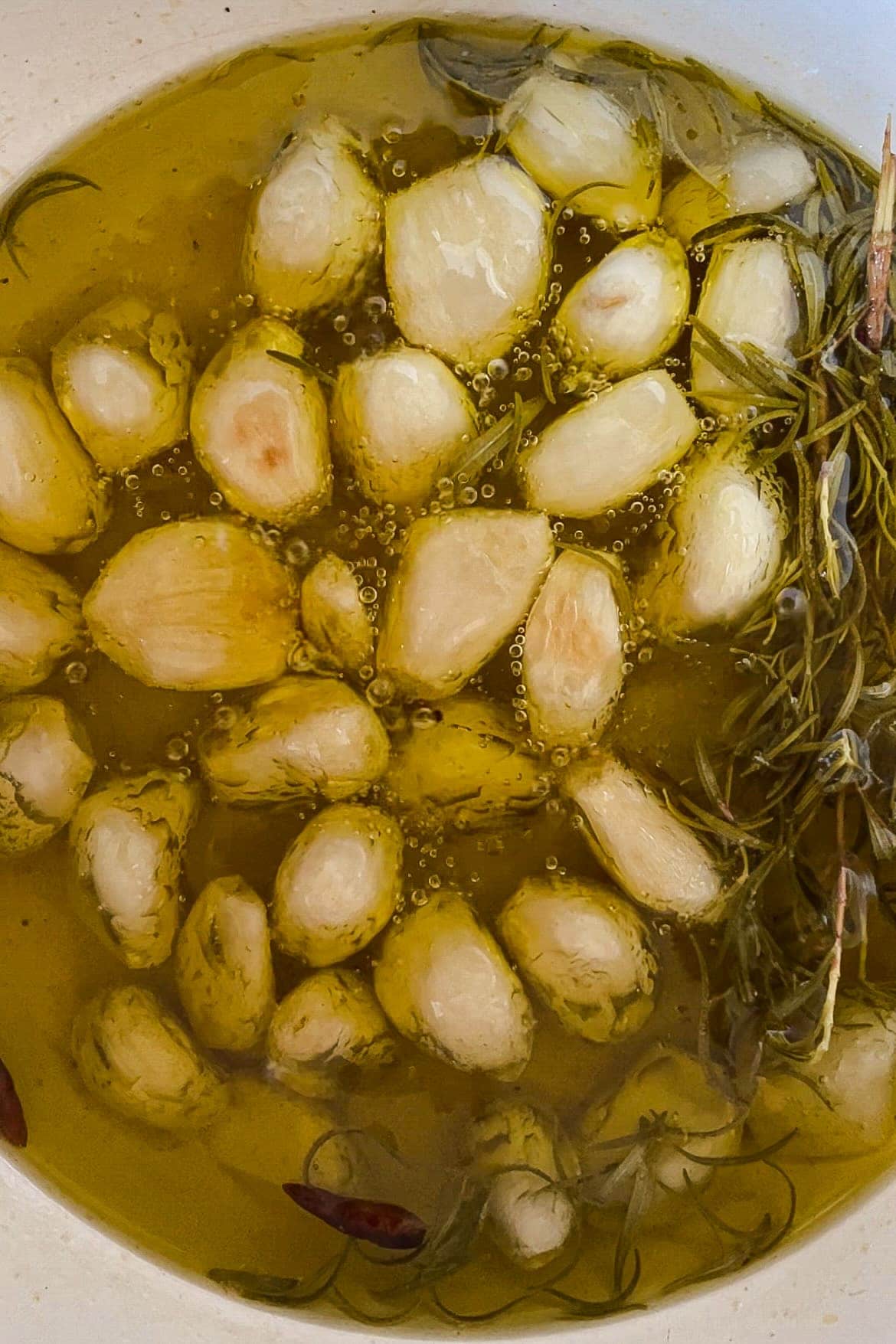 Esta receta de Ajos Confitados (o Confit de Ajo) se va a convertir en tu nuevo indispensable en la cocina porque es tan fácil y los ajos confitados son taaaaaan espectaculares y versátiles. Solo necesitas unas cuantas cabezas de ajo, tus especias y hierbas favoritas, mucho aceite de oliva y 30 minutos. El resultado es un ajo que se deshace como mantequilla, con un sabor dulzón y que puedes usar de mil maneras. Además, como bonus te queda un aceite de ajo insuperable. En esta foto se ven los ajos en la cazuela con romero y 2 guindillas a punto de estar listos. | Receta de Paula's Apron