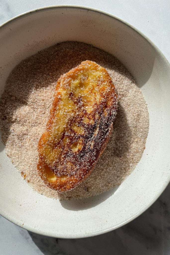 Receta para preparar las mejores Torrijas Tradicionales de leche, caseras, fáciles y quedan tan cremosas. Además, sirven como receta de aprovechamiento para no tirar pan duro. Un postre sencillo y delicioso de Semana Santa. En esta foto se ve una torrija en un plato con la mezcla de azúcar y canela para el rebozado. | Receta de Paula's Apron