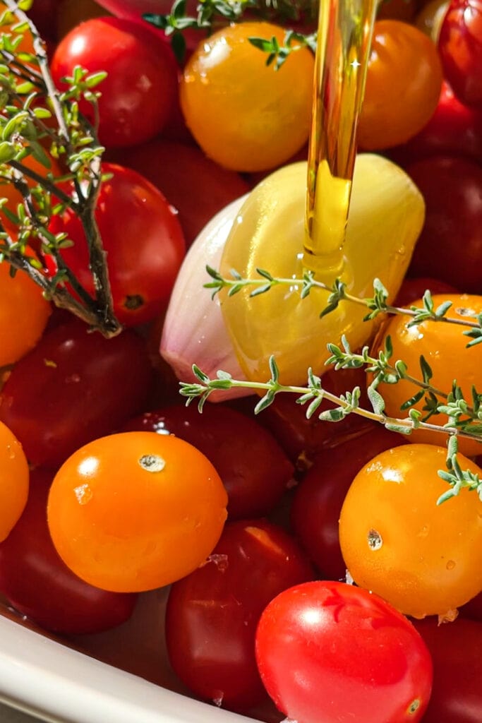 El Tomate Confitado (o Confit de Tomate) es mi forma favorita de conservar el verano. Solo necesitas 5 minutos como máximo de preparación y el resto lo hace el horno. El resultado es un tomate potenciado de sabor, jugoso y dulzón ideal para dar un toque especial y alegrar una infinidad de platos. Lo puedes usar sobre tostadas, con pasta, como topping de untables, carne, pescado, en bocadillos… Hoy le he añadido chalotas, ajo y tomillo pero puedes usar los aromatizantes que más te gusten. En cualquier caso, el aceite infusionado que te va a quedar será delicioso y podrás aprovechar hasta la última gota. En esta foto vemos como se añade aceite para cubrir el tomate en una fuente de horno. | Receta de Paula's Apron