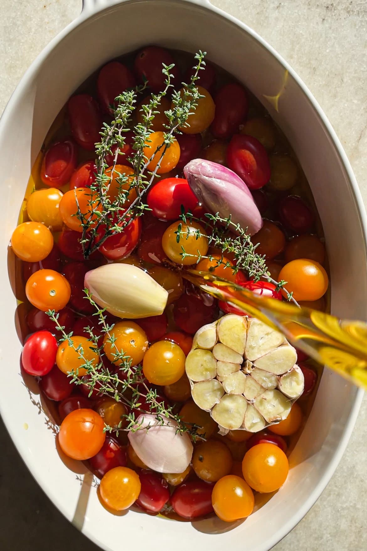 El Tomate Confitado (o Confit de Tomate) es mi forma favorita de conservar el verano. Solo necesitas 5 minutos como máximo de preparación y el resto lo hace el horno. El resultado es un tomate potenciado de sabor, jugoso y dulzón ideal para dar un toque especial y alegrar una infinidad de platos. Lo puedes usar sobre tostadas, con pasta, como topping de untables, carne, pescado, en bocadillos… Hoy le he añadido chalotas, ajo y tomillo pero puedes usar los aromatizantes que más te gusten. En cualquier caso, el aceite infusionado que te va a quedar será delicioso y podrás aprovechar hasta la última gota. En esta foto vemos como se añade aceite para cubrir el tomate en una fuente de horno. | Receta de Paula's Apron