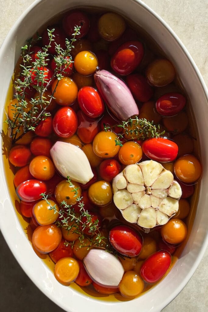 El Tomate Confitado (o Confit de Tomate) es mi forma favorita de conservar el verano. Solo necesitas 5 minutos como máximo de preparación y el resto lo hace el horno. El resultado es un tomate potenciado de sabor, jugoso y dulzón ideal para dar un toque especial y alegrar una infinidad de platos. Lo puedes usar sobre tostadas, con pasta, como topping de untables, carne, pescado, en bocadillos… Hoy le he añadido chalotas, ajo y tomillo pero puedes usar los aromatizantes que más te gusten. En cualquier caso, el aceite infusionado que te va a quedar será delicioso y podrás aprovechar hasta la última gota. Aquí podemos ver todos los ingredientes en una fuente de horno antes de confitarse. | Receta de Paula's Apron