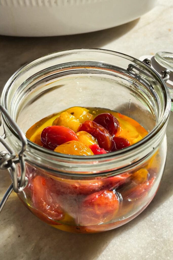 El Tomate Confitado (o Confit de Tomate) es mi forma favorita de conservar el verano. Solo necesitas 5 minutos como máximo de preparación y el resto lo hace el horno. El resultado es un tomate potenciado de sabor, jugoso y dulzón ideal para dar un toque especial y alegrar una infinidad de platos. Lo puedes usar sobre tostadas, con pasta, como topping de untables, carne, pescado, en bocadillos… Hoy le he añadido chalotas, ajo y tomillo pero puedes usar los aromatizantes que más te gusten. En cualquier caso, el aceite infusionado que te va a quedar será delicioso y podrás aprovechar hasta la última gota. | Receta de Paula's Apron