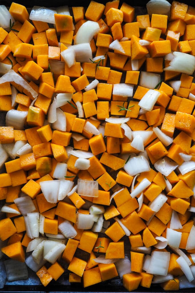 Calabaza y cebolla cortadas en trozos con aceite, sal, pimienta y romero para hacer crema de calabaza. | Receta de Paula's Apron