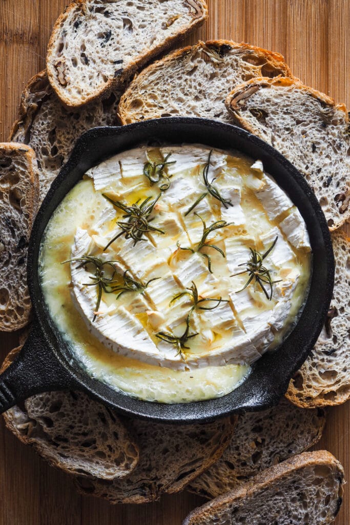 Esta receta de camembert al horno con ajo, romero y nueces caramelizadas es festiva, fácil de preparar y ¡siempre enamora! Rápida, pocos ingredientes y súper deliciosa. El ajo y romero aportan un aroma increíble al Camembert, ya de por si un poco frutal y complejo. Para acompañar, unas tostadas y la guinda del pastel, unas nueces caramelizadas con sirope de arce que también puedes usar para dipear o como acompañamiento de cualquier tabla de quesos y embutidos. Aquí se ve el camembert horneado con el ajo y romero acompañado de tostadas de pan y sin el topping de nueces. | Receta de Paula's Apron