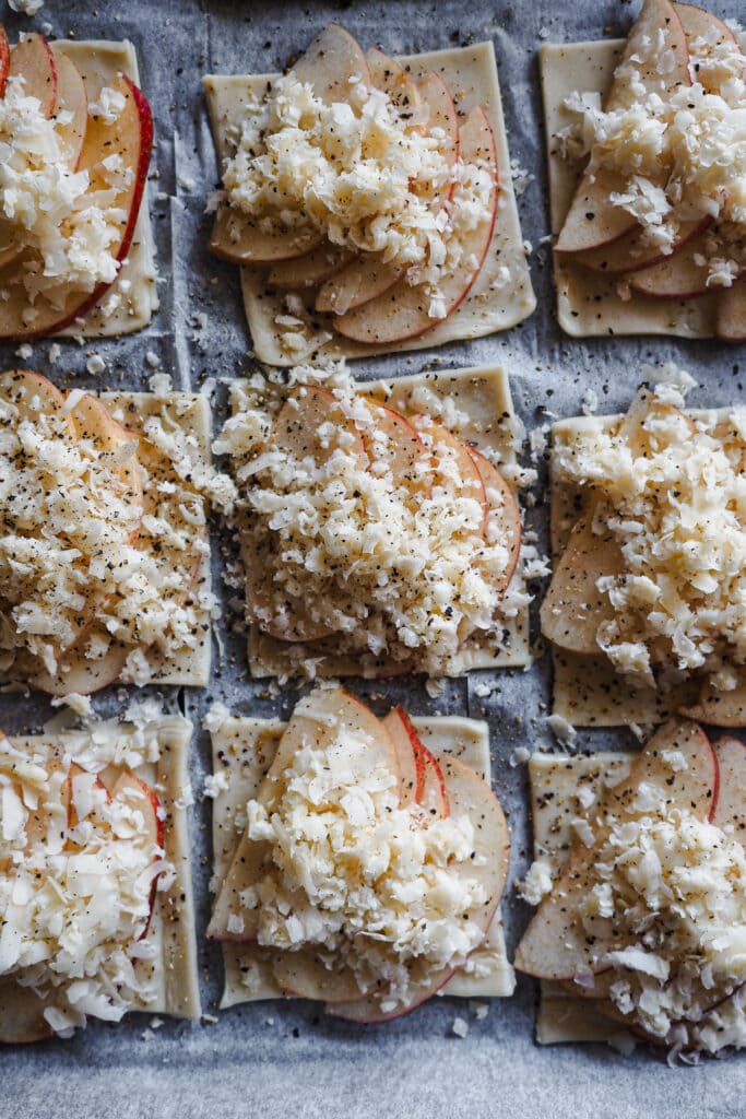 Estos Hojaldritos de Manzana con Panceta y Cheddar son un aperitivo ideal durante otoño e invierno. Mezclan a la perfección dulce y salado, con la manzana de temporada que tan bien le va a la panceta. Y te sorprenderá la combinación, pero un toque de cheddar maduro queda de maravilla, con su punto de sabor un poco salado pero sin ser demasiado fuerte. Son fáciles de hacer y solo llevan 5 ingredientes, ¡simplemente perfectos! En esta foto se ven ensamblados (hojaldre, manzana, queso y pimienta negra) antes de ser horneados. | Receta de Paula's Apron