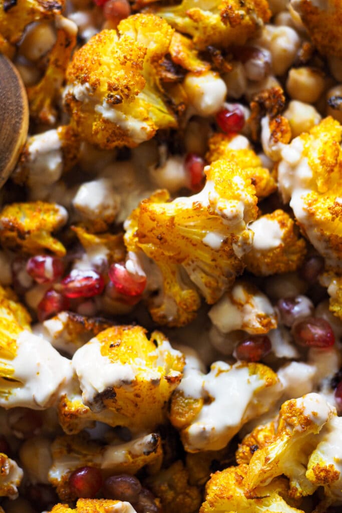 Esta receta de Coliflor Especiada al Horno con Garbanzos y Aliño de Ajo Asado y Tahini te cambiará la forma de ver la coliflor. La coliflor asada queda súper sabrosa, con ese punto tostado y crujiente en su exterior. Y para dar aún más sabor, la aderezo con ajo, pimentón y cúrcuma, que también aportan un toque de color. Para tener un plato más completo nutricionalmente, la mezclamos con garbanzos. Y, como buena ensalada, tiene que tener su aliño, en este caso un aliño cremoso de tahini súper versátil y adictivo. Para terminar, un poco de granada que aporta dulzor, color y crujiente. | Receta de Paula's Apron