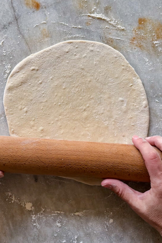 Dando forma a un pan naan con rodillo de madera sobre superficie de mármol enharinada.