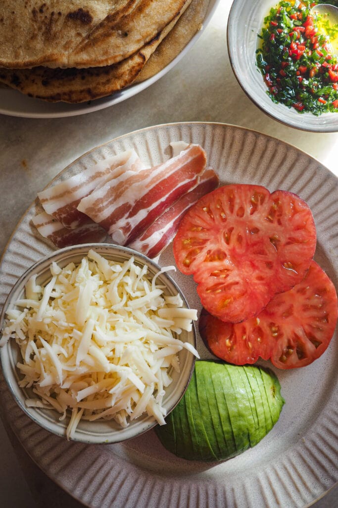 Ingredientes para Bocadillo de Tortilla con Pan Naan, Aguacate, Tomate y Bacon. Lleva pan naan (o cualquier pan plano como roti, tortilla de harina, m'semen o piadina), huevo, queso rallado como mozzarella, provolone, cheddar, emmental, parmesano y/o gruyère, tomate, bacon o panceta, aguacate y un toque de hierbas frescas con chile opcional. | Receta de Paula's Apron