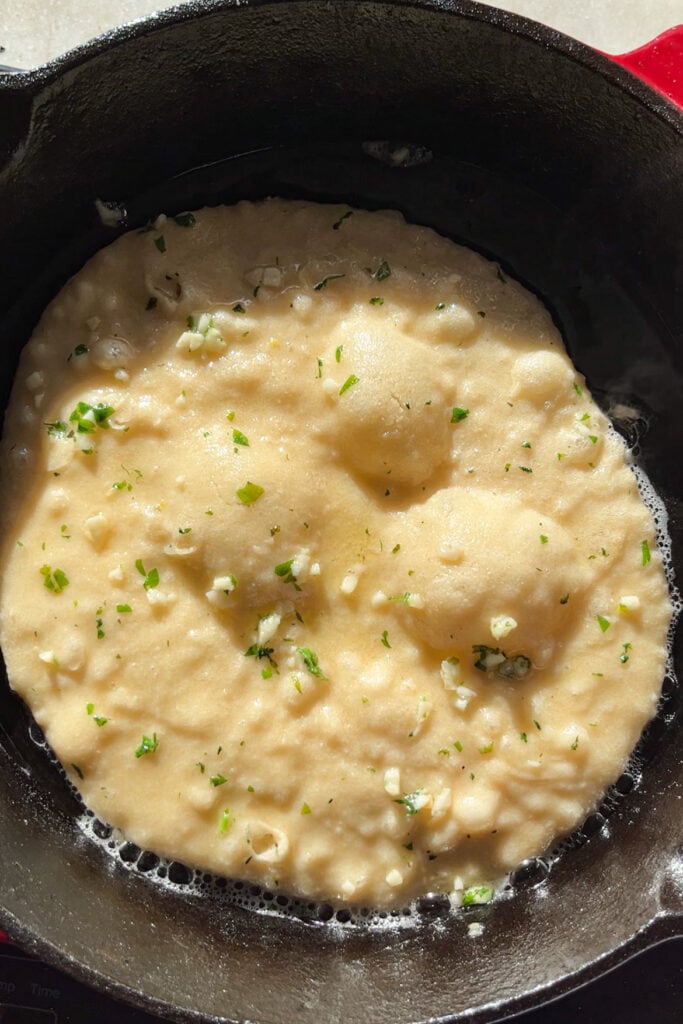 Pan naan cociéndose en una sartén con la parte superior aún sin hacer, llena de burbujas y pincelada con mantequilla de ajo y perejil.