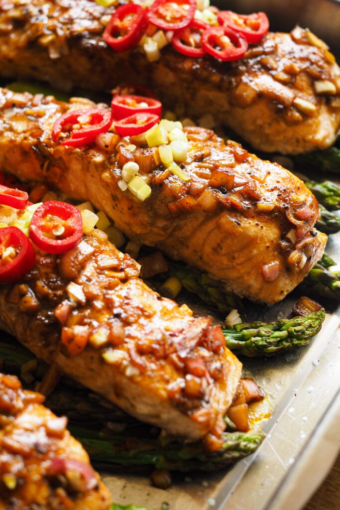 Este Salmón al Horno con Espárragos y Vinagreta de Miel y Limón es fresco, ligero, fácil de hacer y nutritivo, ¡lo tiene todo! El salmón se marina con una vinagreta deliciosa que mezcla un toque ácido con dulce y salado. Luego se cocina todo junto en una bandeja de horno y terminamos con un topping de chile y cebolleta. Sírvelo con arroz o puré de patatas y tienes un plato completo para una comida saludable, o acompáñalo de ensalada si quieres algo más light para la cena. ¡Una delicia! Aquí vemos 4 filetes de salmón sobre una cama de espárragos ya horneados, la carne del salmón se puede ver que ya no está cruda, con topping de chile y cebolleta a falta de un golpe de grill. | Receta de Paula's Apron