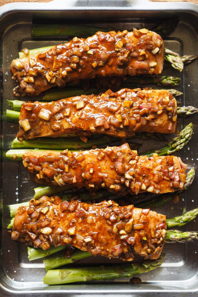 Este Salmón al Horno con Espárragos y Vinagreta de Miel y Limón es fresco, ligero, fácil de hacer y nutritivo, ¡lo tiene todo! El salmón se marina con una vinagreta deliciosa que mezcla un toque ácido con dulce y salado. Luego se cocina todo junto en una bandeja de horno y terminamos con un topping de chile y cebolleta. Sírvelo con arroz o puré de patatas y tienes un plato completo para una comida saludable, o acompáñalo de ensalada si quieres algo más light para la cena. ¡Una delicia! Aquí vemos 4 filetes de salmón marinados encima de una cama de espárragos en una bandeja de horno antes de ser horneados. | Receta de Paula's Apron
