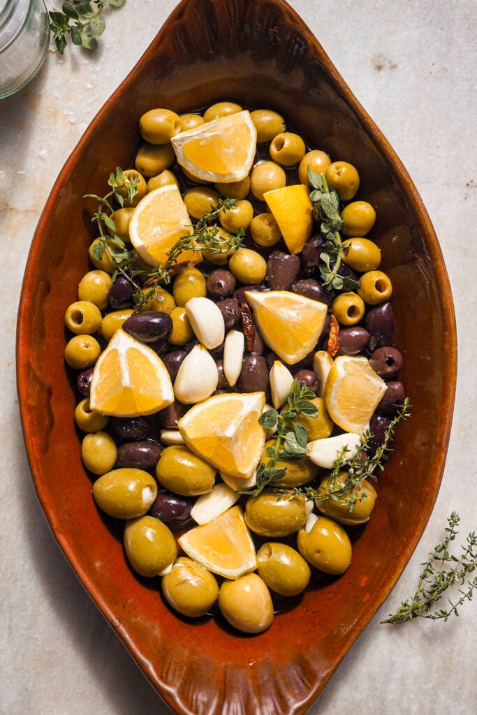 Estas Aceitunas Asadas con Burrata son un aperitivo facilísimo y que siempre impresiona. Solo necesitas 5 minutos como máximo de preparación y el resto lo hace el horno. Quedan unas aceitunas aromáticas y el toque final de burrata... Simplemente es la guinda del pastel, aunque puedes omitirla y usar las aceitunas para mejorar una infinidad de platos. Eso sí, si las sirves como aperitivo, acompáñalas de un buen pan. Aquí las puedes ver en una fuente de horno antes de asarse con hierbas, limón, dientes de ajo y guindillas. | Receta de Paula's Apron