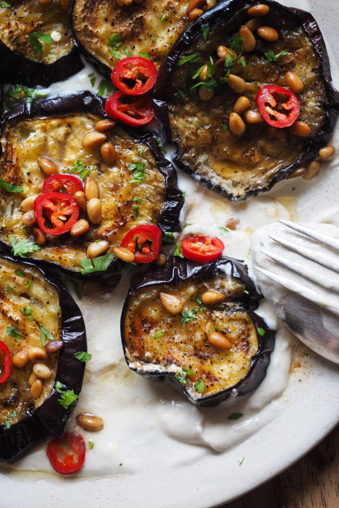 Esta Berenjena Asada con Salsa de Yogur y Tahini se deshace en boca, y con el contraste de la salsa de yogur y tahini queda aún más bestial. El sabor asado de la berenjena, con la frescura de la salsa, del perejil y el chile fresco (opcional), y el toque crujiente de los piñones con su mantequilla tostada es pura explosión de todos los sentidos y en todos los sentidos. Disfrútala como aperitivo o acompañamiento y si tienes algún reticente de la berenjena a tu alrededor, hazle cambiar de opinión con este recetón. | Receta de Paula's Apron