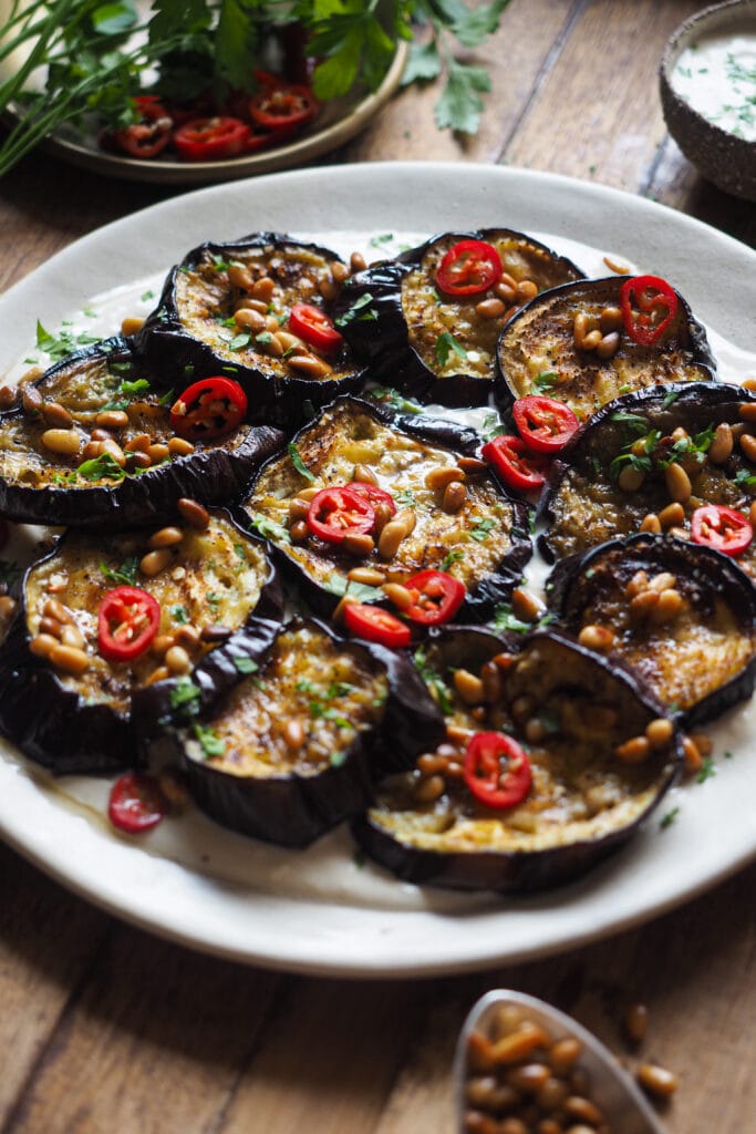 Esta Berenjena Asada con Salsa de Yogur y Tahini se deshace en boca, y con el contraste de la salsa de yogur y tahini queda aún más bestial. El sabor asado de la berenjena, con la frescura de la salsa, del perejil y el chile fresco (opcional), y el toque crujiente de los piñones con su mantequilla tostada es pura explosión de todos los sentidos y en todos los sentidos. Disfrútala como aperitivo o acompañamiento y si tienes algún reticente de la berenjena a tu alrededor, hazle cambiar de opinión con este recetón. | Receta de Paula's Apron