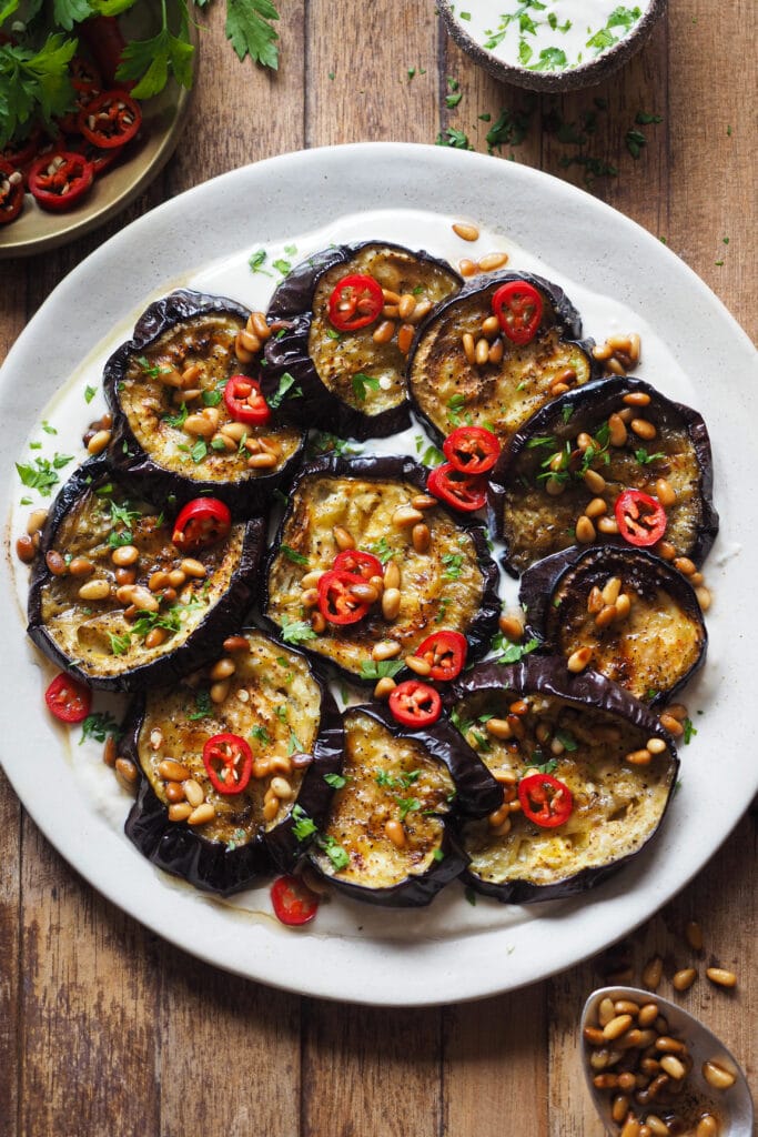 Esta Berenjena Asada con Salsa de Yogur y Tahini se deshace en boca, y con el contraste de la salsa de yogur y tahini queda aún más bestial. El sabor asado de la berenjena, con la frescura de la salsa, del perejil y el chile fresco (opcional), y el toque crujiente de los piñones con su mantequilla tostada es pura explosión de todos los sentidos y en todos los sentidos. Disfrútala como aperitivo o acompañamiento y si tienes algún reticente de la berenjena a tu alrededor, hazle cambiar de opinión con este recetón. | Receta de Paula's Apron