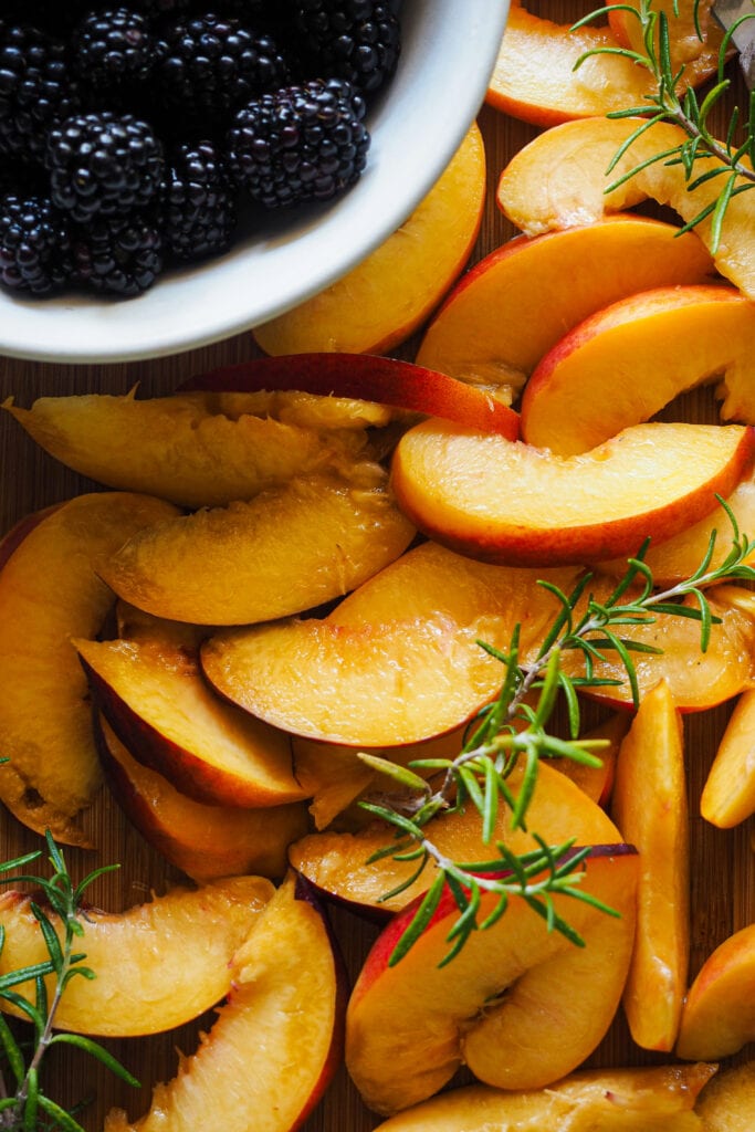 Nectarinas cortadas a gajos, moras en un bol y ramitas de romero sobre un tabla de madera.