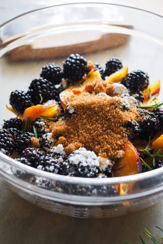 Relleno Galette de Nectarina y Moras al Romero: en un bol de cristal hay nectarina cortada a gajos delgados, moras, almidón de maíz, azúcar moreno, vainilla y hojas de romero.