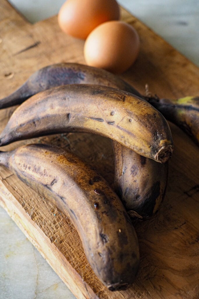 3 plátanos maduros con piel y 2 huevos sobre un tabla de madera para hacer banana bread o bizcocho de plátano.