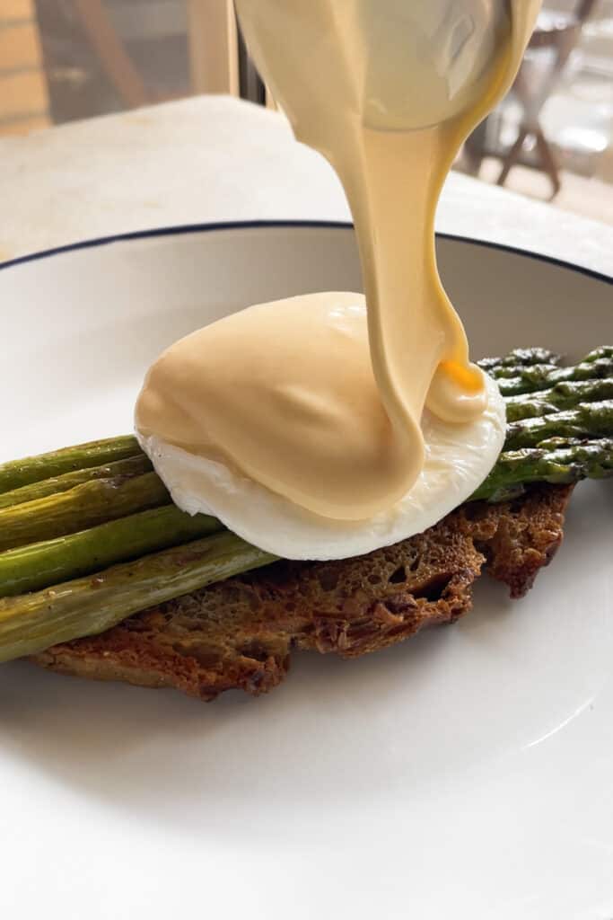 Virtiendo salsa holandesa sobre un huevo escalfado que a su vez está sobre una tostada con espárragos trigueros salteados. | Receta de Paula's Apron