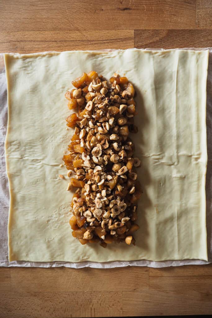 Paso 2 montar trenza de hojaldre rellena de manzana: avellanas tostadas y troceadas sobre manzana caramelizada que a su vez está en el centro y a lo largo de una lámina de hojaldre.