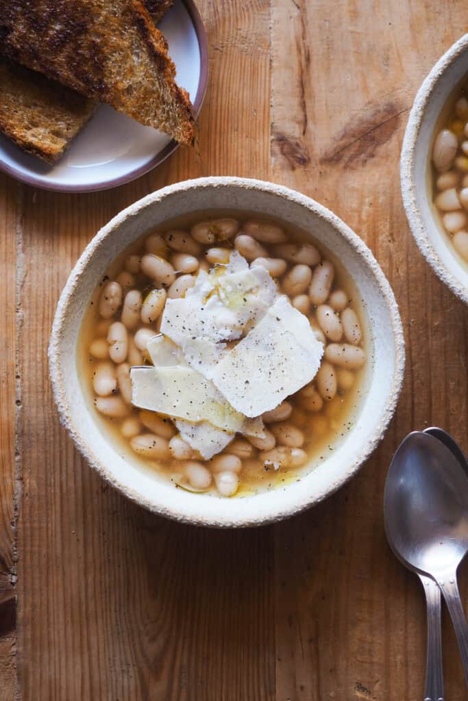 Cómo cocer alubias es algo que todos deberíamos saber hacer. Además de rebote te sale un caldo sabroso, nutritivo, fácil y versátil que puedes usar de mil formas, así como las alubias cocidas resultantes. Las alubias secas son mucho más económicas que comprarlas en bote, puedes hacer en cantidad y conservarlas para usarlas en una infinidad de platos, como esta sopa de alubias y parmesano. | Receta de Paula's Apron