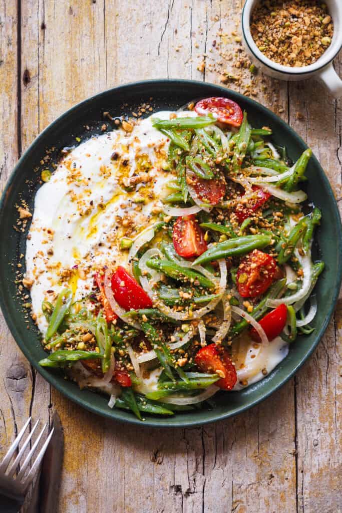 Esta Ensalada de Judías Verdes y Tomate con Dukkah es una versión reinventada de un clásico de mi infancia. No sé tu, pero yo he crecido comiendo ensalada de judías verdes, tomate y cebolla con atún (y a veces huevo duro) cada verano de mi vida, semana sí semana también XD. Hoy mantengo las hortalizas y la vinagreta de mostaza que siempre hacía mi abuela para aliñarla, un combo sencillo, pero perfecto. Y le añado un toque crujiente con Dukkah casero, que además le aporta un punto aromático, junto con una base de yogur que lo redondea todo con su cremosidad y ligera acidez. En resumen, una ensalada fresquita perfecta para esta temporada. | Receta de Paula's Apron