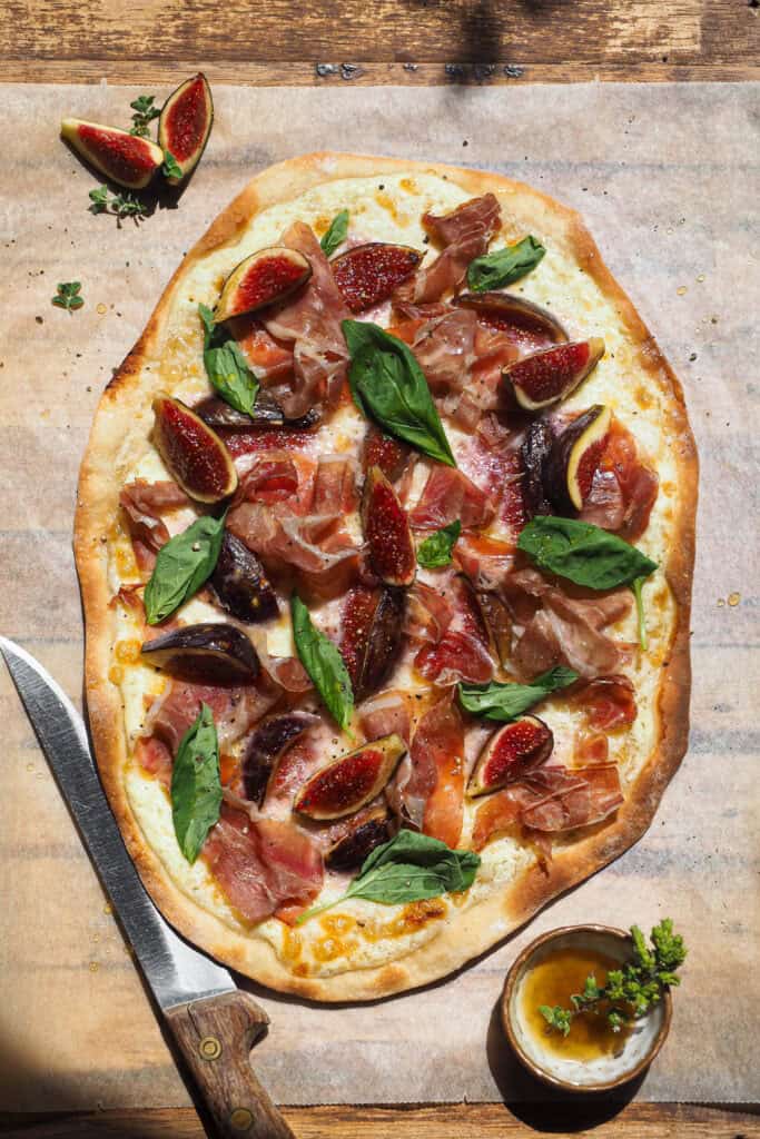 Esta versión de Flammkuchen de Higos y Jamón (Tarte Flambée de Higos y Jamón) es perfecta para finales de verano, cuando da menos pereza encender el horno y los higos están de temporada. Lleva queso y jamón para aportar contraste de color y ese equilibrio perfecto entre dulce y salado. El resultado es una pizza fina, crujiente, ligera y muy sabrosa que siempre deja a todo el mundo queriendo repetir. | Receta de Paula's Apron