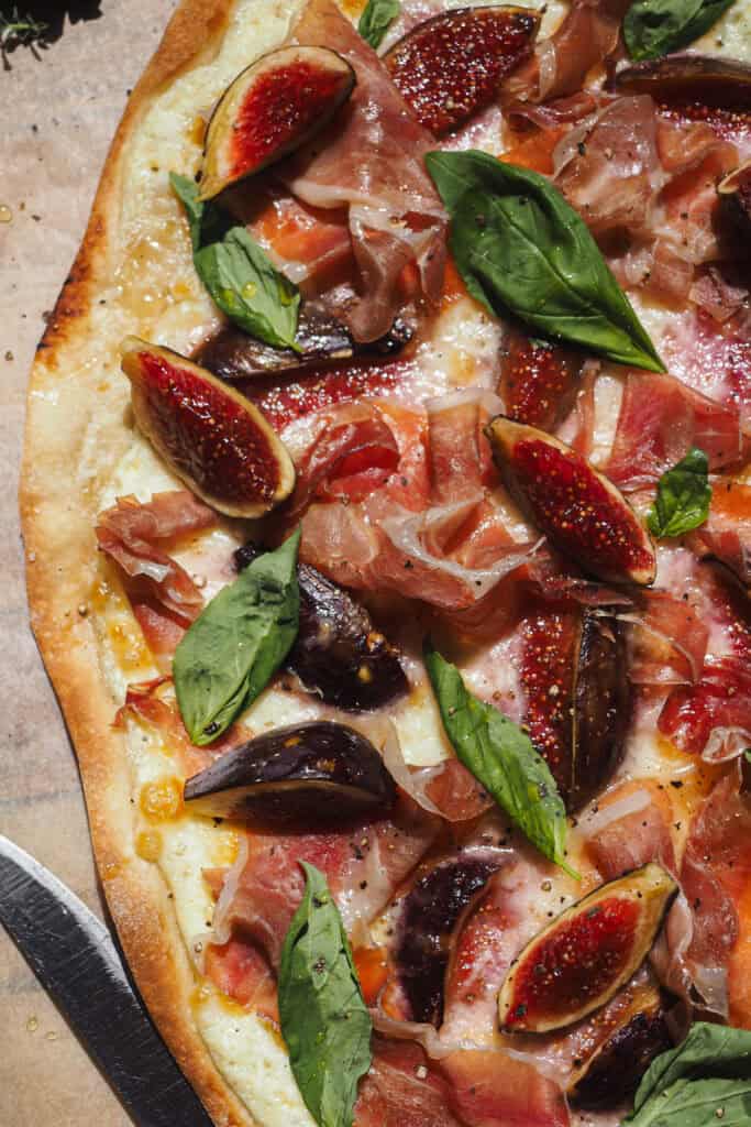 Esta versión de Flammkuchen de Higos y Jamón (Tarte Flambée de Higos y Jamón) es perfecta para finales de verano, cuando da menos pereza encender el horno y los higos están de temporada. Lleva queso y jamón para aportar contraste de color y ese equilibrio perfecto entre dulce y salado. El resultado es una pizza fina, crujiente, ligera y muy sabrosa que siempre deja a todo el mundo queriendo repetir. | Receta de Paula's Apron