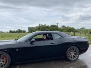 Black muscle car with a person smiling in the driver's seat, scenic countryside background with green fields and trees, overcast sky, vehicle featuring a sleek design and sporty rims, ideal for car and pet fashion enthusiasts.