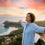 A person standing on a mountain overlooking a scenic coastline at sunset with arms wide open, representing the feeling of freedom and success.