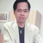 Benedict in a light-colored suit with a cross necklace, indoor setting.