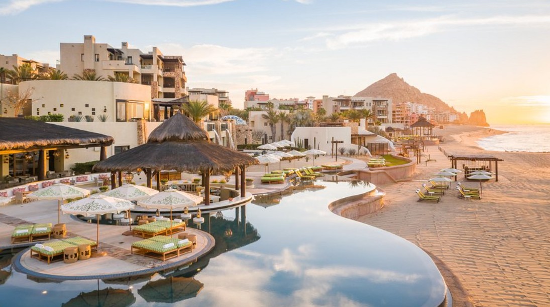 Scenic Sunrise Over A Luxury Beachfront Resort With Palapas, Loungers, And Mountain Views In Pedregal Real Estate, Cabo San Lucas.