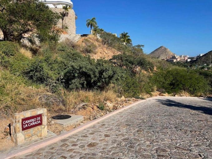 Cobblestone Street With Lush Greenery And Hillside Homes In Pedregal, Cabo San Lucas, Mexico, Featuring Mountain Views And Clear Blue Sky, Showcasing Prime Pedregal Real Estate Locations.