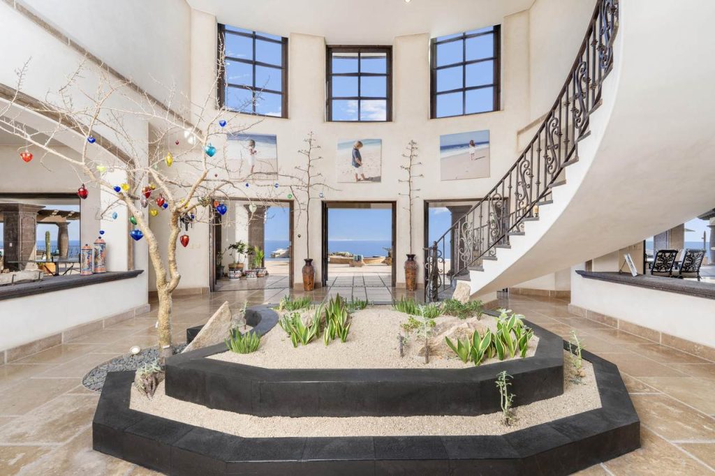 Elegant foyer at Casa Encantada #100 in Pedregal, Cabo San Lucas, featuring a curved staircase, indoor garden with desert landscaping, high windows, and ocean views in the background.