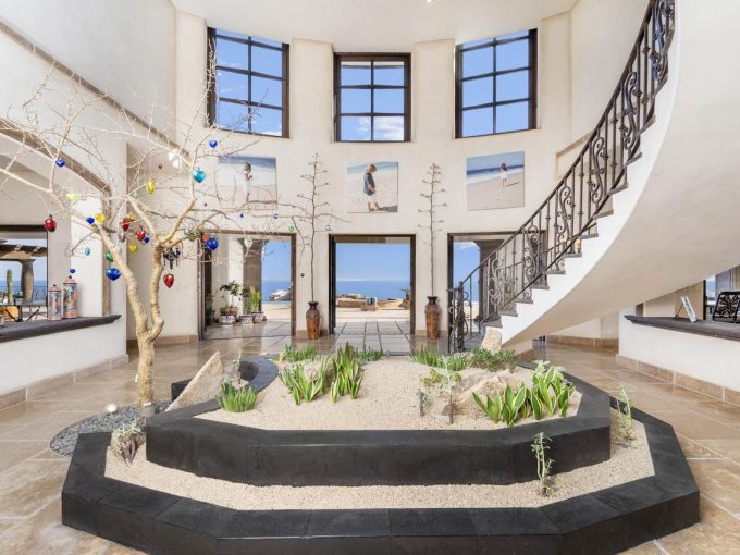 Elegant Foyer At Casa Encantada #100 In Pedregal, Cabo San Lucas, Featuring A Curved Staircase, Indoor Garden With Desert Landscaping, High Windows, And Ocean Views In The Background.