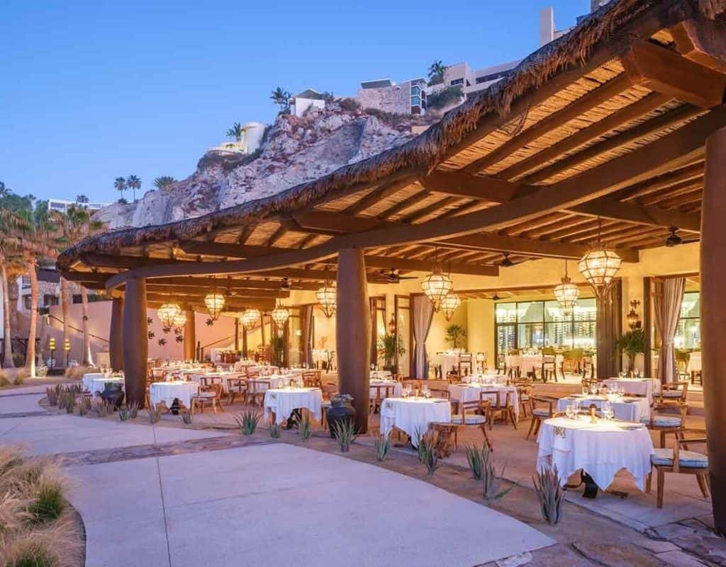 Elegant Outdoor Dining Area At Pedregal With White Tablecloths, Wooden Chairs, And Warm Lighting Under A Thatched Roof, Offering A Luxurious Dining Experience.