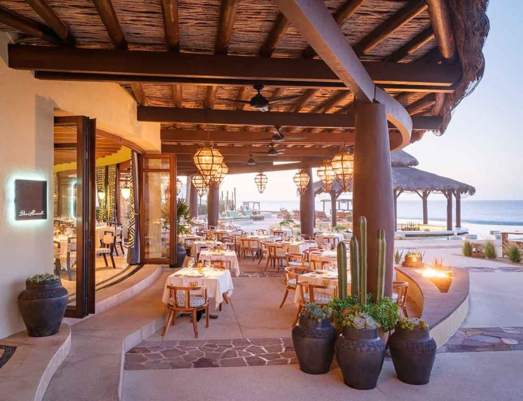 Luxurious Open-Air Beachfront Restaurant At Pedregal, Featuring Elegant Tables, Warm Lighting, And Stunning Ocean Views, Perfect For A Memorable Dining Experience.